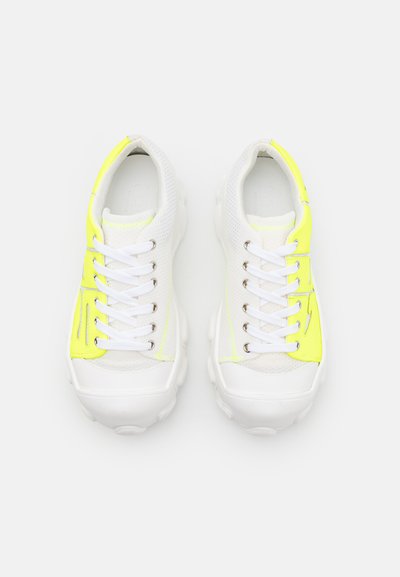 Λευκά αθλητικά παπούτσια από δίχτυ με φωτεινές κίτρινες λεπτομέρειες, στρογγυλή μύτη, παχιά σόλα και λευκά κορδόνια. Υφή στην επάνω επιφάνεια με ελαφριά γυαλάδα και λεπτομέρειες ραφής.