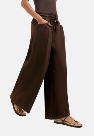 Person iført mørkebrune, vidde bukser med forlommer og sorte thong-sandaler, stående mod en ensfarvet baggrund.