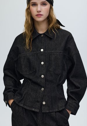 Svart jeansjacka med silverknappar, två bröstfickor och en avslappnad passform. Har en krage och sänkta axlar.