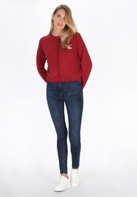 Sarkans, garām piedurknēm, kardigans ar pogu aizdari un ribotu apakšmalu, kombinēts ar tumši zilām šaurām džinsām un baltām kedām.