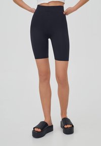Femme portant un short cycliste taille haute noir et des sandales plateformes noires, debout devant un fond clair uni.