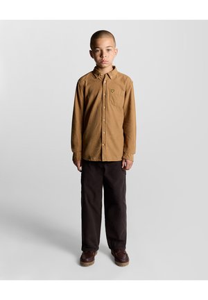 Junge steht frontal zur Kamera, trägt ein beigefarbenes Hemd mit Knöpfen, dunkle Hosen und braune Schuhe vor einem einfarbigen hellen Hintergrund.