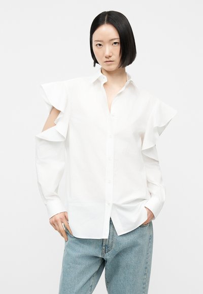 Femeie cu bob negru drept, purtând o bluză albă cu mâneci lungi și decupaje stil „cold-shoulder” cu volane, precum și blugi albastre deschis, stând cu mâinile în buzunare.