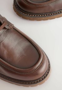 Zapatos de cuero marrón con punta redondeada, que presentan una textura suave, detalles de costura y una suela de goma de color marrón claro.