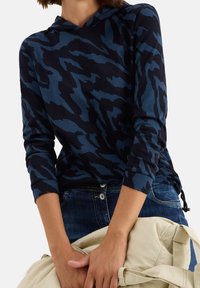 Sweat à capuche bleu marine avec un motif de rayures de tigre en bleu plus foncé, doté de manches longues et d'une capuche avec cordon de serrage. Porté avec un jean foncé, tenant un sac clair.