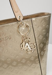 Sac à main Guess ton doré avec logos embossés, porte-clés décoratif doré avec "LA" et charms de marque supplémentaires, et anse beige.