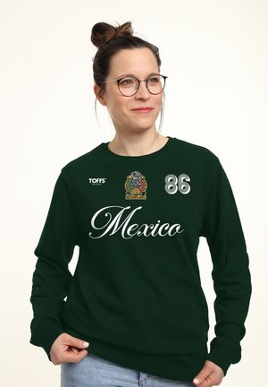 Frau mit Brille und hochgebundenen Haaren, die einen dunkelgrünen Mexico-Sweatshirt mit Logo, Nummer 86 und dem Schriftzug „TOFFS“ trägt und vor weißem Hintergrund posiert.