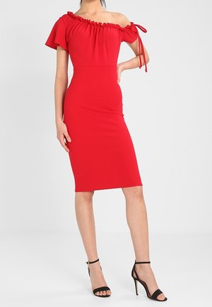 Vrouw draagt een nauwsluitende rode jurk met blote schouders, ruches en korte mouwen, gecombineerd met zwarte sandalen met hoge hakken.