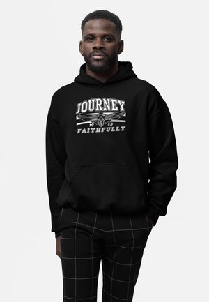 Mand i sort hætte sweatshirt med teksten "Journey Faithfully 1983" og emblem, parret med sorte bukser med hvide rutenetlinjer.