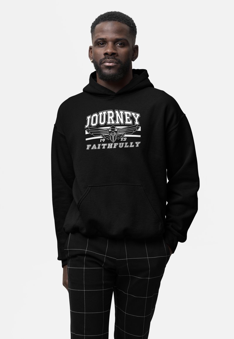 Mand i sort hætte sweatshirt med teksten "Journey Faithfully 1983" og emblem, parret med sorte bukser med hvide rutenetlinjer.