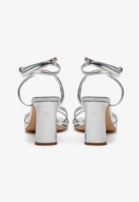 Silver heeled sandals with narrow crisscross straps, block heels, and metallic finish. Smooth texture with a rounded toe and open back design.