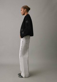 Suéter negro texturizado con patrones de estrellas en las mangas, combinado con jeans de tiro alto en gris claro y zapatos negros de punta. La modelo está de lado.