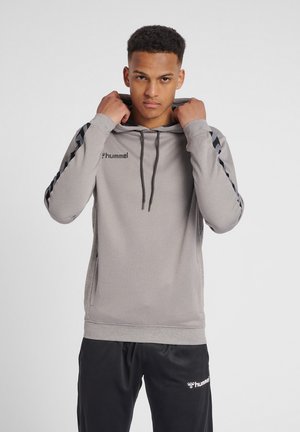 Giovane che sistema il cappuccio di una felpa grigia hummel, indossa pantaloni sportivi neri hummel, in piedi davanti a uno sfondo semplice.