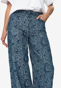 Jeans med vid ben som har en mörkblå tyg med ett blommigt ros-mönster i ljusare blått. Inkluderar midjeresår och sidofickor.