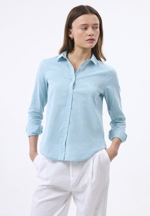 Kobieta ubrana w jasnoniebieską koszulę na guziki z podwiniętymi rękawami i białe spodnie, stoi z rękami w kieszeniach, patrząc w bok.