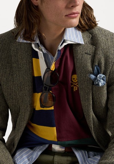 Tweed blazer, lagd med en stripet skjorte og en flerfarget rugbyshirt; solbriller henger fra kragen; teksturerte og mønstrede stoffer synlige.