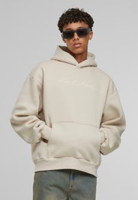 Beige hættebluse med frontlomme og broderet logo i hvid. Materialet ser blødt ud med en afslappet pasform og ribbede manchetter.