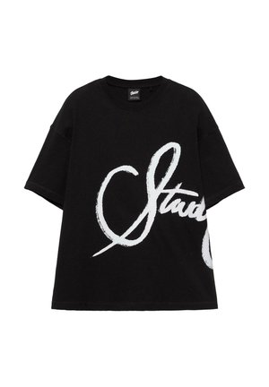 T-shirt noir à manches courtes avec une grande écriture cursive blanche partiellement épelée "Staud" sur le devant.