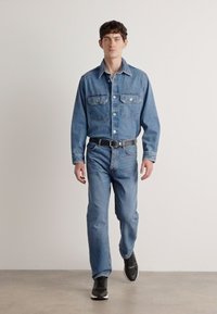 Chemise en denim avec col classique, deux poches poitrine et fermetures à bouton ; associée à un jean en denim clair et des chaussures de sport noires.