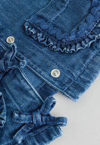 Tessuto di denim con una sfumatura azzurro chiaro, caratterizzato da bordi arricciati, rifiniture a zigzag e bottoni a pressione argentati. I dettagli includono fiocchi annodati sulle maniche.