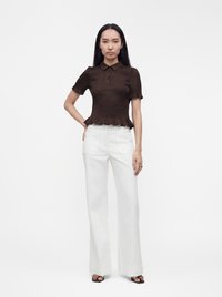 Femme portant un haut texturé marron foncé à manches courtes et un pantalon blanc taille haute à jambes larges, debout avec une main sur la hanche.