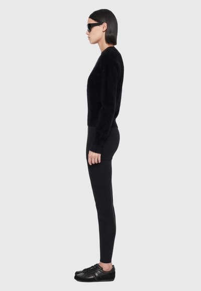 Une femme se tient de profil, portant des lunettes de soleil noires, un pull noir, un legging et des baskets sur un fond gris clair.
