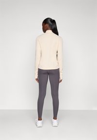 Beige langermet topp paret med mørkegrå leggings, med en tettsittende design og tommelhull, samt en glatt og elastisk tekstur. Hvite Sneakers.