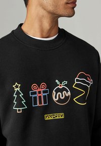 Sudadera negra con gráficos bordados coloridos: un árbol de Navidad, una caja de regalo, un pudín navideño y un icono del personaje Pac-Man, todos contorneados.
