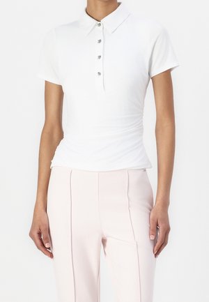 Person trägt ein enganliegendes weißes Poloshirt mit kurzen Ärmeln und vier Knöpfen sowie eine hellrosa Hose mit hoher Taille und steht vor einem schlichten Hintergrund.