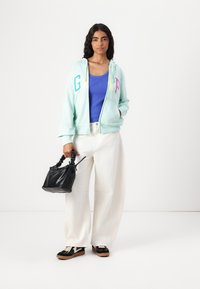 Mintgrøn zip-up hoodie med multicolor "GAP" bogstaver, båret over en blå ribbet top. Sammen med hvide brede bukser og en sort taske.