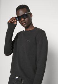 Maglione nero a costine con scollatura rotonda e una piccola etichetta con logo, abbinato a occhiali da sole neri oversize.