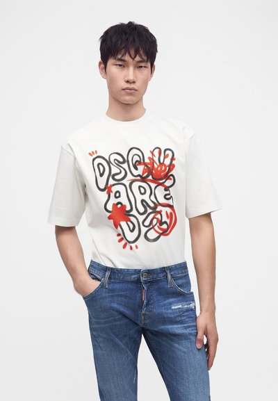 Jeune homme portant un t-shirt graphique blanc avec un texte abstrait noir et rouge et un jean bleu, debout avec une main dans la poche.