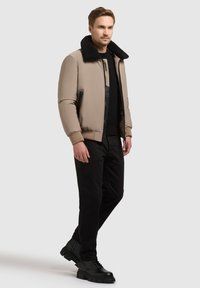 Veste bomber beige avec un col en fourrure noire, poche zippée, poignets côtelés, portée sur un pull noir et un pantalon noir, accompagnée de bottes noires.