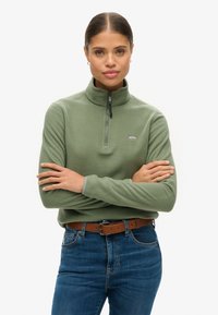 Groene fleecetopper met hoge kraag en halve ritssluiting, met een klein logo op de borst. Gecombineerd met blauwe jeans en een bruine riem.