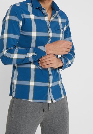 Homme portant une chemise à carreaux bleue et blanche boutonnée avec un pantalon gris à cordon, ajustant sa manche gauche avec sa main droite.