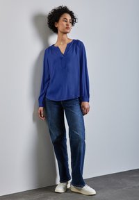 Blusa azul de manga larga con escote en V y hombros fruncidos, combinada con jeans de mezclilla oscura y zapatillas blancas, todo sobre un fondo neutro.