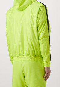 Neon grøn træningsjakke med hætte, elastisk talje og manchetter. Har en sort stribe langs ærmerne og en blank finish.