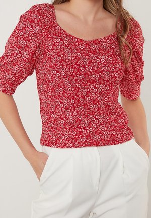 Vrouw draagt een rode top met bloemenpatroon en pofmouwen, witte broek met hoge taille, handen in de zakken, tegen een neutrale achtergrond.