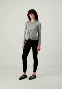 Camisola cinza de manga longa com detalhe torcido à frente, combinada com leggings pretas justas. Modelo usa sapatos slip-on pretos, de pé contra um fundo neutro.