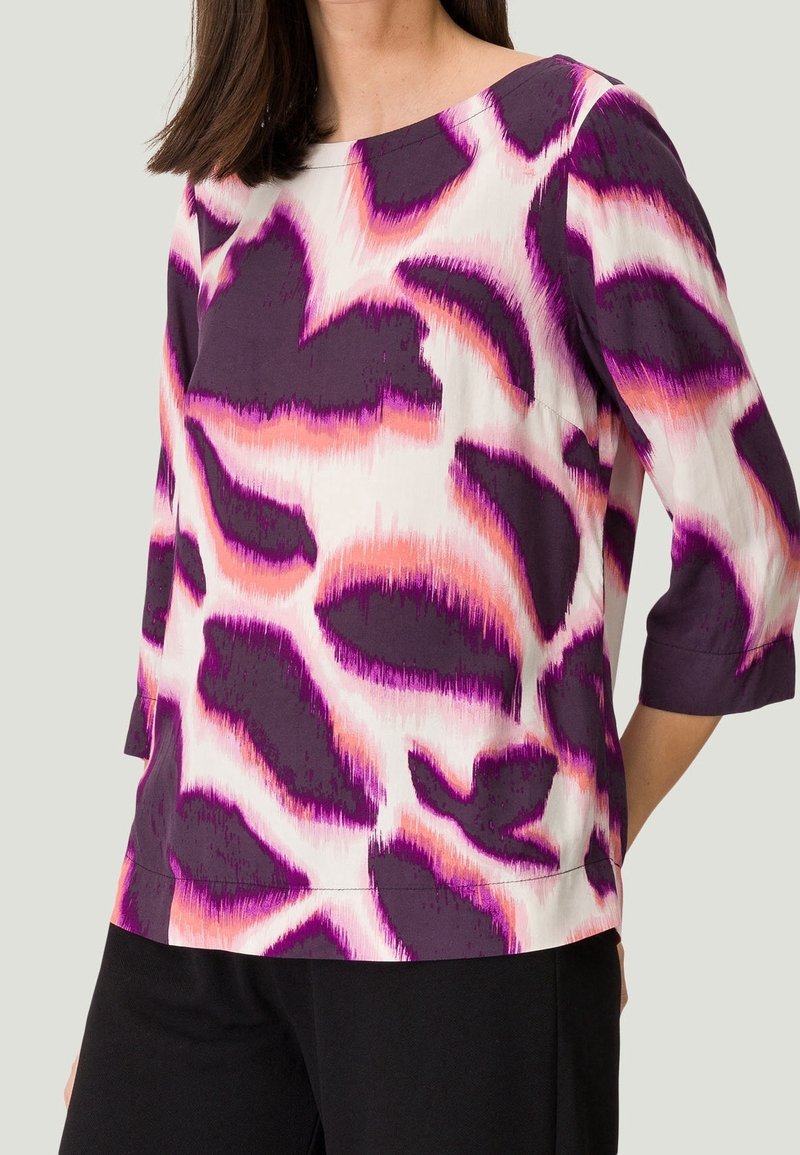 Lila und weiße Bluse mit auffälligen abstrakten Mustern in Pink und Lila. Mit Dreiviertelärmeln und rundem Halsausschnitt. Glatte Stoffoberfläche.