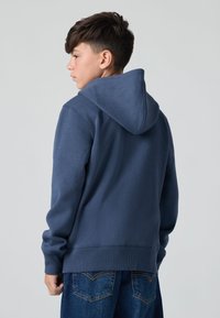 Felpa blu navy con cappuccio regolabile, polsini e orlo a costine, abbinata a jeans in denim scuro. Il tessuto ha una consistenza liscia.