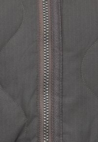 Hall kangaga, millel on ruuduline muster, on keskel tõmblukk. Tõmblukul on metallhammastega ja must tõmbelint, mida ümbritseb kontrastne õmblus.