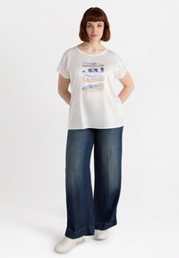Camiseta blanca de manga corta con un diseño de estampado apilado en azul y beige, combinada con pantalones de mezclilla oscuros de pierna ancha y zapatillas blancas.