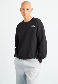 Plikas žmogus, apsirengęs juodu „The North Face“ sportiniu megztiniu ir šviesiais pilkais „The North Face“ sportiniais kelnais, stovi su rankomis kišenėse.
