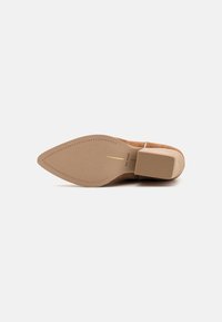 Zapato de tacón alto beige con un tacón cuadrado y punta afilada. La suela es lisa y presenta un pequeño patrón de relieve, además de un acento dorado.