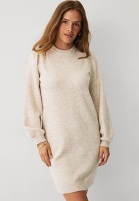 Ung kvinde iført en lysebeige langærmet strikket sweaterkjole med opkrøllede ærmer, stående mod en ensfarvet hvid baggrund.