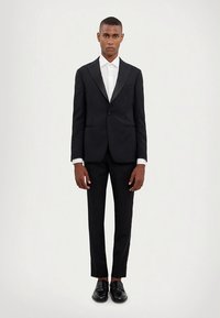 Zwart op maat gemaakt pak met een slanke pasvorm, voorzien van een eenknopsluiting, revers met inkeping en broek met platte voorkant; gecombineerd met een wit overhemd.