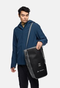 Man in blauw jack die een zwarte en grijze Vaude schoudertas met een voorste gesp sluiting vasthoudt en naar de tas kijkt.