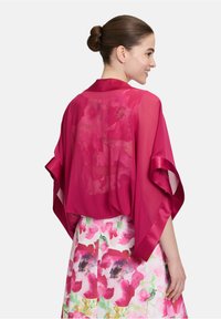 Transparenter magentafarbener Kimono mit weiten Ärmeln, der eine glatte Textur aufweist. Getragen über einem rock mit floralem Muster in kräftigen Farben und Designs.