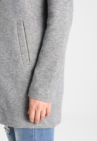 Cappotto grigio in lana con una finitura testurizzata, dotato di una tasca laterale e di una manica dritta. Abbinato a jeans chiari strappati.